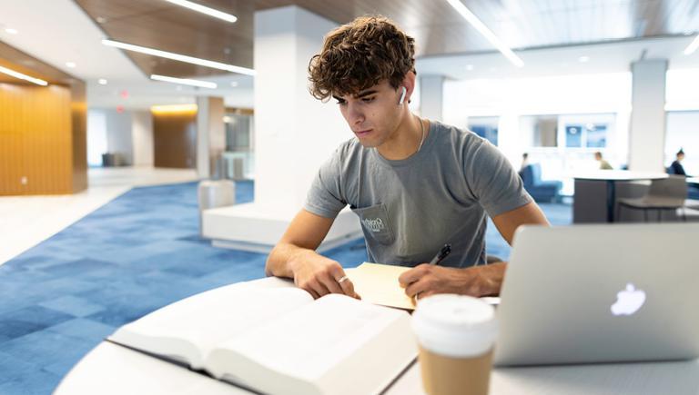 Creighton student studying at table with laptop, books and notes.